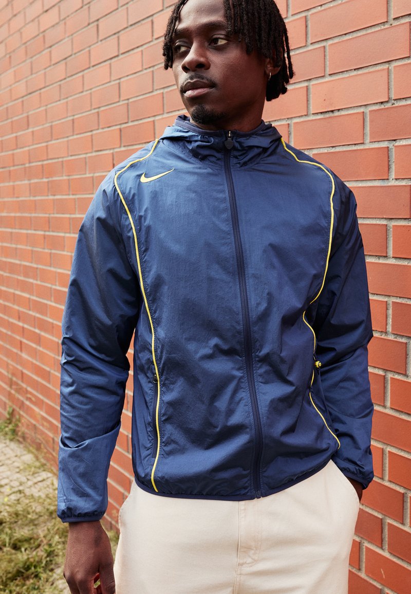 Chaqueta tipo rompevientos de la marina con detalles en amarillo, textura ligera, capucha y cierre con cremallera, combinada con pantalones beige. Fondo de pared de ladrillo.