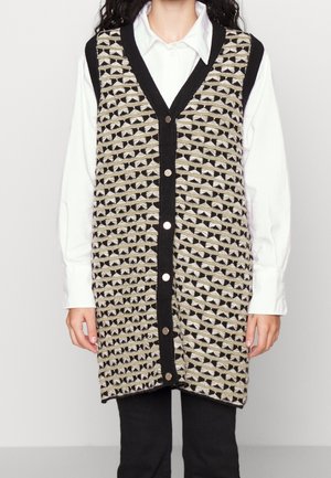 Kobieta ubrana w długi czarno-beżowy, wzorzysty, zapinany na guziki dziergany kamizelka na białą koszulę z kołnierzykiem i czarne spodnie, stojąca na jednolitym tle.