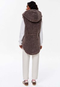 Hnědý vestník z umělé ovčí srsti s velkou kapucí, dlouhým zadním designem a bočními kapsami, nošený přes bílou mikinu a krémové kalhoty.