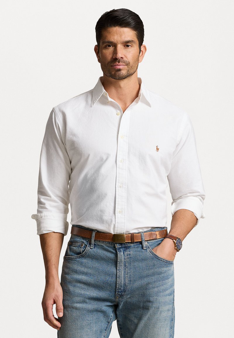 Chemise blanche à manches roulées, en coton, associée à un jean en denim bleu et une ceinture marron, avec un logo sur la chemise.