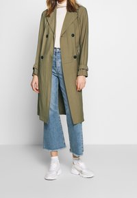 Kobieta ubrana w oliwkowy trencz zapinany na podwójną brodę, biały golf, przetarte niebieskie dżinsy, białe skarpetki i białe trampki.