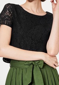 Top in pizzo nero con scollatura ampia e maniche corte, abbinato a una gonna verde a vita alta con pieghe e dettaglio della cintura annodata.