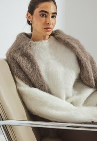 Kremowy i brązowy, puszysty sweter z luźnymi rękawami, założony pod miękki brązowy szalik. Model siedzi na nowoczesnym, eleganckim krześle.