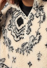 Polarowy sweter z ćwierć zamkiem, w kolorze kremowym z czarnymi abstrakcyjnymi wzorami. Miękka tekstura z wyraźnymi detalami przeszycia.