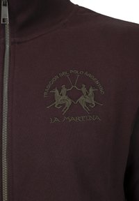 Burgundy zip-up jacket made of cotton blend, featuring an embossed logo and two polo players graphic in olive green. Smooth texture.