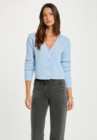 Hellblauer gestrickter Cardigan mit V-Ausschnitt und langen Ärmeln, ausgestattet mit silbernen herzförmigen Knöpfen und einem cropped Design. Getragen mit grauen Jeans.