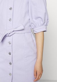 Vestido lilás com botões, de mangas curtas bufantes, cintura ajustada e cinto de laço próprio. Apresenta detalhes em botões prateados e um tecido de textura suave.