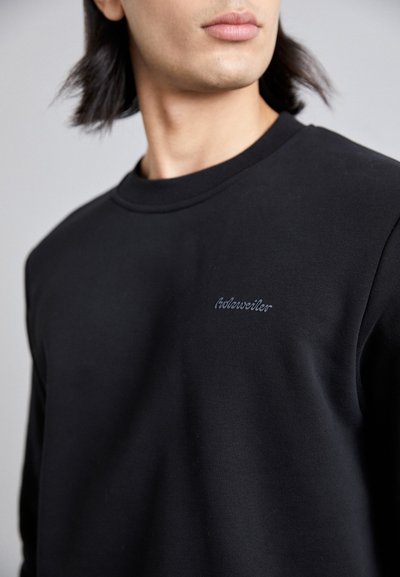 Sort bomulds sweatshirt med rund hals og subtil broderet branding på brystet. Glat tekstur og afslappet pasform.