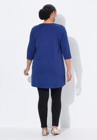 Blaue Tunika mit Dreiviertelärmeln, kombiniert mit schwarzen Leggings und flachen Metallic-Schuhen, zeichnet sich durch einen geraden Schnitt und seitliche Schlitze aus.