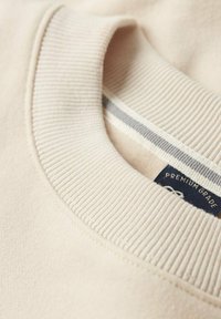 Sudadera color beige claro con un cuello acanalado y una textura suave. Presenta una pequeña etiqueta azul oscura con "GRADO PREMIUM" y un detalle a rayas en el interior del cuello.