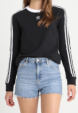 Personne portant un t-shirt noir Adidas à manches longues avec des bandes blanches sur les manches et un short en jean bleu clair taille haute.