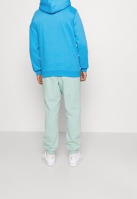 Blå hoodie med känguruficka, kombinerad med myntgrön träningsoverall och vita sneakers med gulddetaljer. Enfärgad bakgrund.