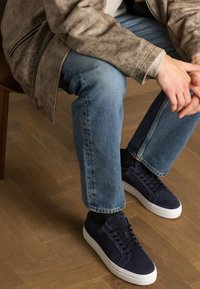Sneakers in suede blu navy con lacci neri, suola in gomma bianca, indossati con jeans in denim azzurro chiaro e una giacca in pelle marrone consumata, seduto su un pavimento in legno.