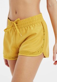 Geel zwemshorts met een elastische tailleband en zijkoord. Heeft een kantachtige uitsnijpatroon langs de zoom en de zijkanten.