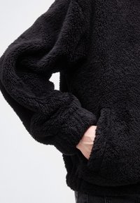 Άτομο που φοράει μαύρο, αφράτο φλις μπουφάν με το ένα χέρι μέσα σε μπροστινή τσέπη, σε απλό ανοιχτόχρωμο φόντο.