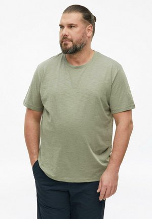 Mann mit Bart und zurückgebundenem Haar, trägt ein grünes Kurzarm-T-Shirt und dunkle Hose, steht mit einer Hand in der Tasche und schaut nach links.