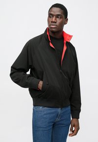 Sort jakke med høj krave og røde detaljer, der har lynlåslukning, sidelommer og ribstrikkede manchetter. Bært over en sort t-shirt.