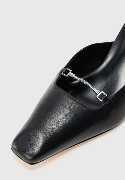 Scarpa di pelle nera con punta affusolata, texture liscia e un elemento intagliato con hardware metallico che esalta il design.