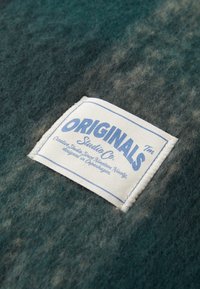 Un tejido de lana verde texturizado con tonos variables, que presenta una etiqueta cosida en blanco que dice "ORIGINALS Studio Co" en azul.
