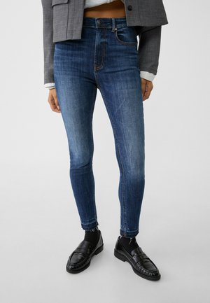 Femme portant un jean skinny bleu foncé avec des ourlets effilochés, des chaussettes noires, des mocassins noirs, une veste courte grise et un haut blanc.