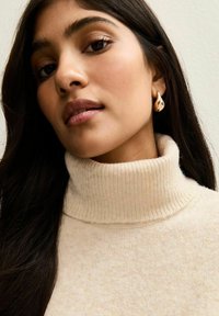 Crèmekleurige coltrui van gebreid materiaal, met een geribbeld kraagdetail. Draagt gouden druppelvormige oorbellen. Glad, steil donker haar.