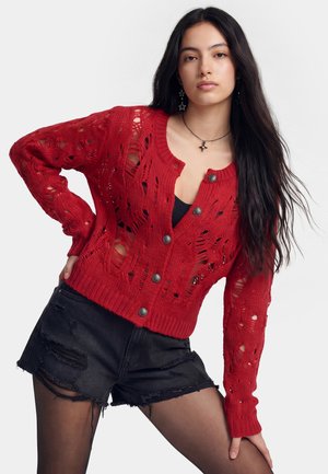 Ung kvinde med langt mørkt hår iført en rød slidt strikket cardigan, sort tanktop, sorte revnede shorts og stjerneformede øreringe og halskæde.