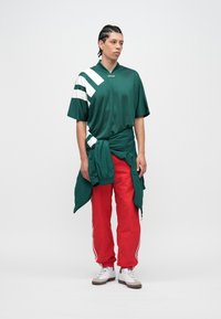 Groen-wit T-shirt met korte mouwen en een V-hals, gecombineerd met rode broek met elastische taille en witte strepen. Witte schoenen met bruine accenten.