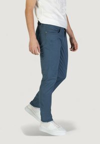 Pantalones elásticos azules de corte ajustado y superficie texturizada, con múltiples bolsillos y una cremallera, combinados con zapatillas blancas.