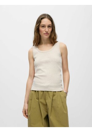 Femme aux longs cheveux bruns portant un haut beige sans manches et un pantalon ample vert olive, debout devant un fond blanc uni.