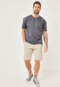 Graues Baumwoll-T-Shirt mit kurzen Ärmeln, kombiniert mit hellbeigen Shorts und grauen Sneakers. Das Model hat Tattoos auf seinen Armen.
