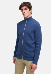 Blå teksturert strikket zip-up genser med stående krage og kontrastdetaljer på glidelåsen, kombinert med beige bukser.