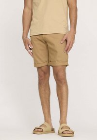 T-shirt en coton beige, associé à un short marron clair avec des revers, et des sandales marron clair à deux brides.