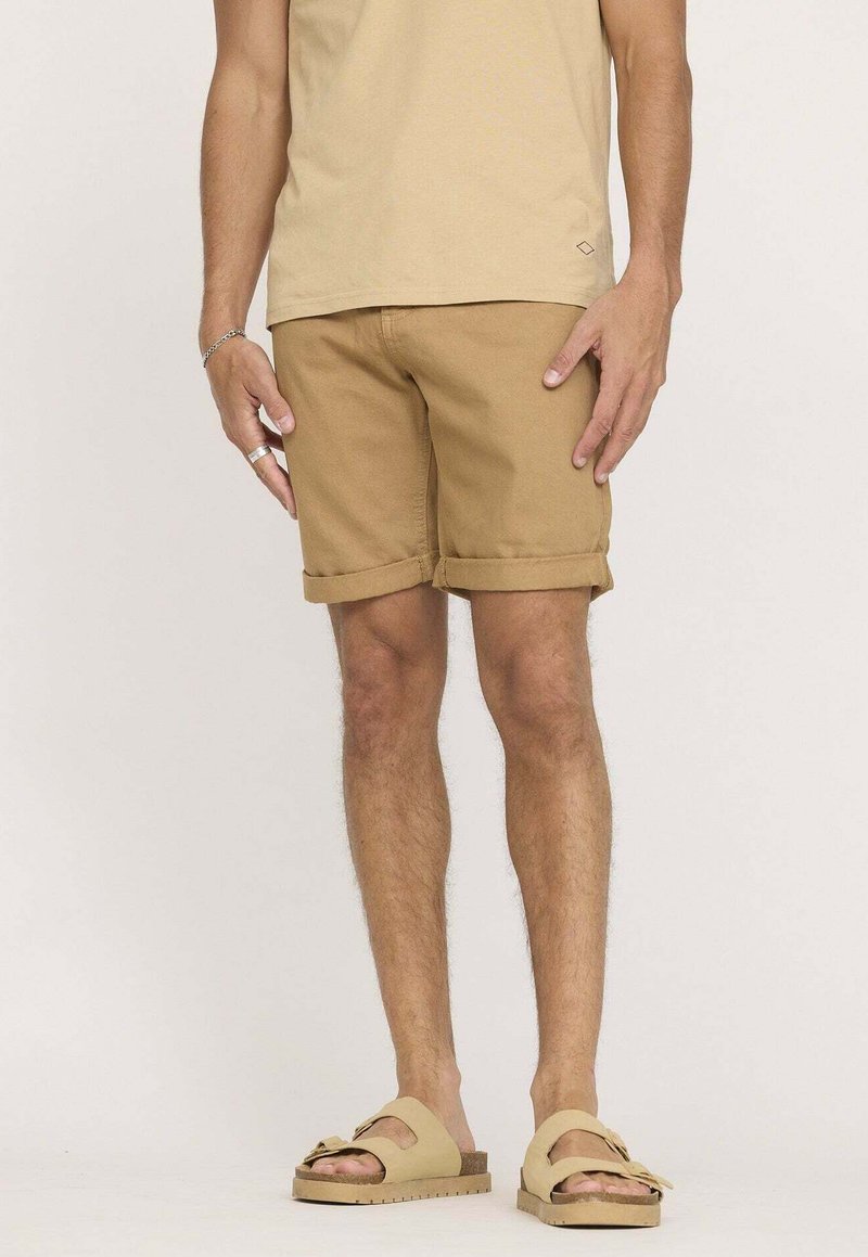 T-shirt en coton beige, associé à un short marron clair avec des revers, et des sandales marron clair à deux brides.