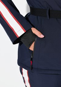 Námořnická modrá bunda se bílými a červenými pruhy na rukávech, s černým elastickým pásem a kapsou na zip s červeným táhlem.