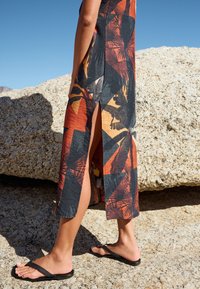 Abito lungo con audace motivo geometrico in nero, arancione e tonalità terrose; realizzato in tessuto leggero con spacco laterale, abbinato a infradito nere.