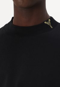 Sort bomulds t-shirt med ribbet rund hals. Har et lille guldlogo broderet på skulderen. Glat tekstur, tætsiddende design.