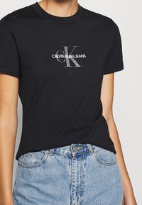 Osoba nosząca czarną koszulkę z dżinsowej kolekcji Calvin Klein oraz jasnoniebieskie dżinsy z wysokim stanem, pokazana od szyi do bioder.