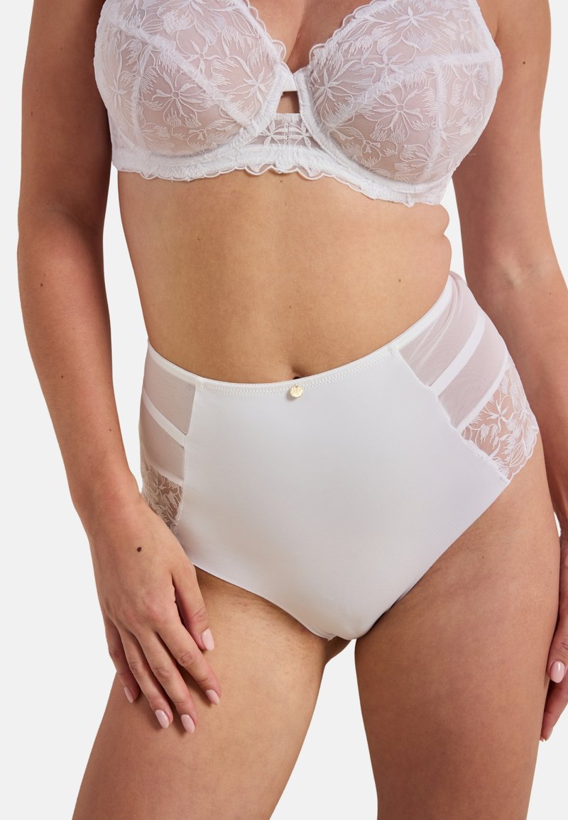 Femme portant un soutien-gorge en dentelle blanche et une culotte taille haute blanche avec des panneaux latéraux en dentelle et en maille, sur un fond uni.