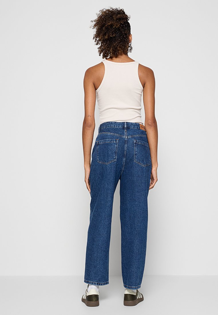 Frau mit lockigem Haar, die ein weißes ärmelloses Oberteil, blaue hoch geschnittene Jeans, weiße Socken und dunkle Sneakers trägt und vor einer schlichten Wand steht.
