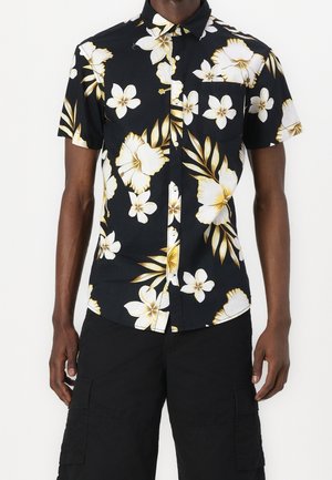 Mężczyzna ubrany w czarną, krótki rękaw, zapinaną na guziki koszulę z dużym białym i żółtym kwiatowym wzorem oraz czarne szorty typu cargo, stojący z luźno opuszczonymi ramionami.