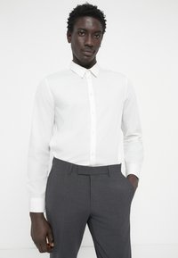 Bela srajca z dolgimi rokavi in gumbi, slim fit, v kombinaciji s sivimi formalnimi hlačami, ki se odlikujejo po mehki teksturi in diskretnih vzorcih.
