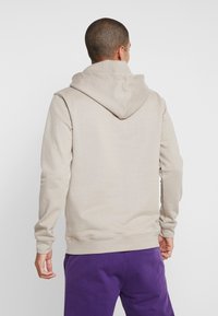 Muž s krátkými vlasy, oblečený v světle béžové mikině s kapucí a fialových kalhotách, stojící zády k fotoaparátu na jednobarevném bílém pozadí.