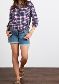 Camisa de cuadros abotonada en tonos azul marino, rosa y blanco; combinada con pantalones cortos de denim con los puños enrollados. El modelo lleva sandalias marrones. Fondo de madera.