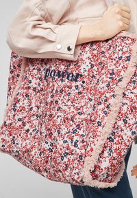 Osoba trzymająca dużą torbę w kwiatowy wzór z różowymi frędzlami i napisem "power." wyhaftowanym na froncie niebieską nitką.