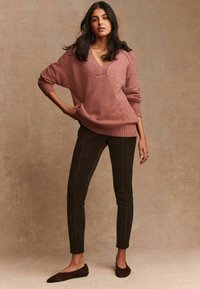Žena u ružičastom oversized V-izrez džemperu, tamnim uskim hlačama i tamnosmešnim baletankama, stoji s jednom rukom na kuku.