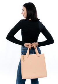 Donna con top nero e jeans che tiene una grande borsa beige dietro la schiena su uno sfondo bianco.