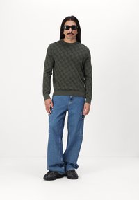 Olivgrüner gestrickter Pullover mit geometrischem Muster; kombiniert mit hellblauen weit geschnittenen Jeans und schwarzen Sneakers. Das Model trägt dunkle Sonnenbrillen.