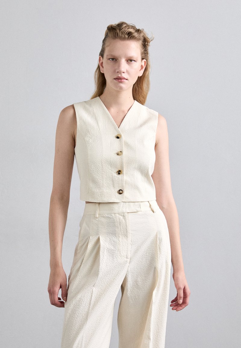 Top sans manches texturé blanc avec un décolleté en V et trois boutons décoratifs, associé à un pantalon large plissé taille haute assorti.