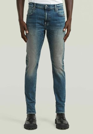 Jean slim bleu pour homme avec des détails délavés porté avec des bottes noires à lacets sur fond uni.