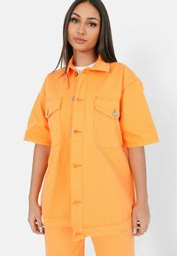 Camisa de mezclilla naranja, de mangas cortas, con cierre de botones, dos bolsillos en el pecho, ajuste relajado, textura suave y detalles de costura en contraste.
